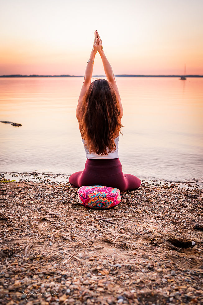 Eine Frau mit langen braunen Haaren sitzt auf einem bunt gemusterten Meditationskissen an einem Kiesstrand im Sonnenuntergang am See