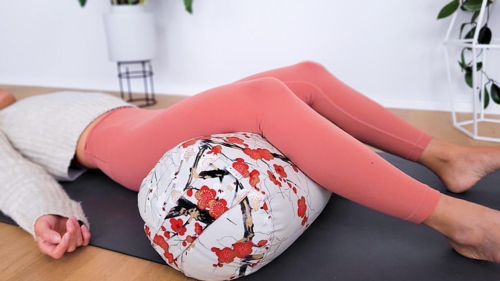 Eine Frau liegt auf einer Yogamatte und übt die Yoga Schlussentspannung mit einem Yogabolster.