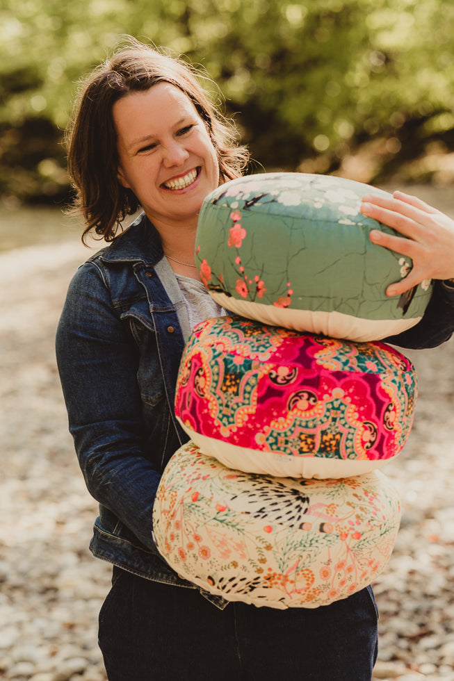 Frau mit blauer Jeansjacke trägt drei verschiedene, bunte Meditationskissen und lacht.