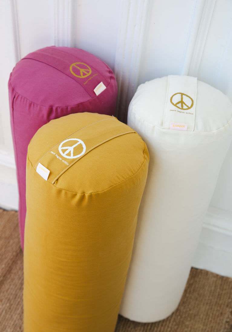 Drei verschiedenen Yoga Bolster in weiß, gelb und pink stehen vor einer weißen Wand.