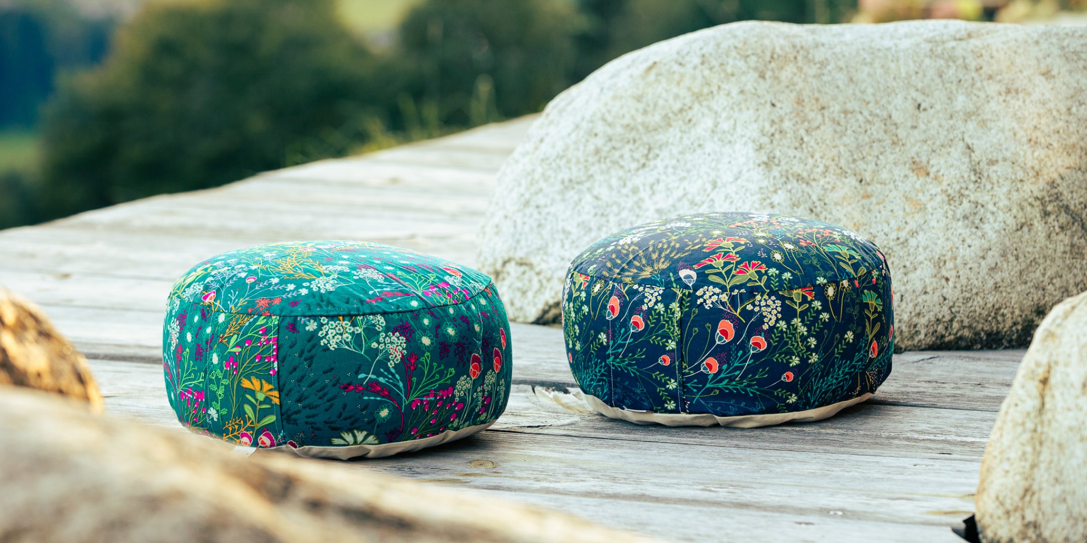 Ein grünes und eine blaues Meditationskissen liegen auf einem Holzdeck. Die Kissen sind mit bunten Blumen bedruckt.