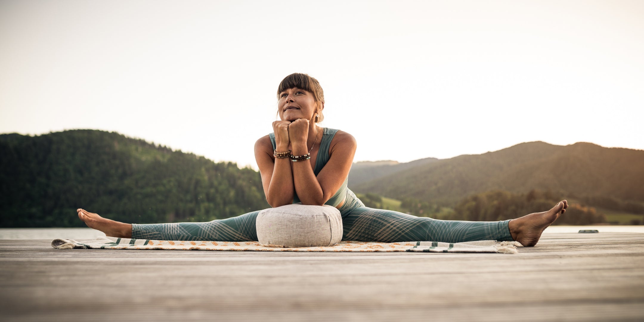 Auf einem Holzdeck sitzt eine Frau in der Grätsche und stützt ihre Ellenbogen auf ein Meditationskissen von Goldwerk Schliersee.