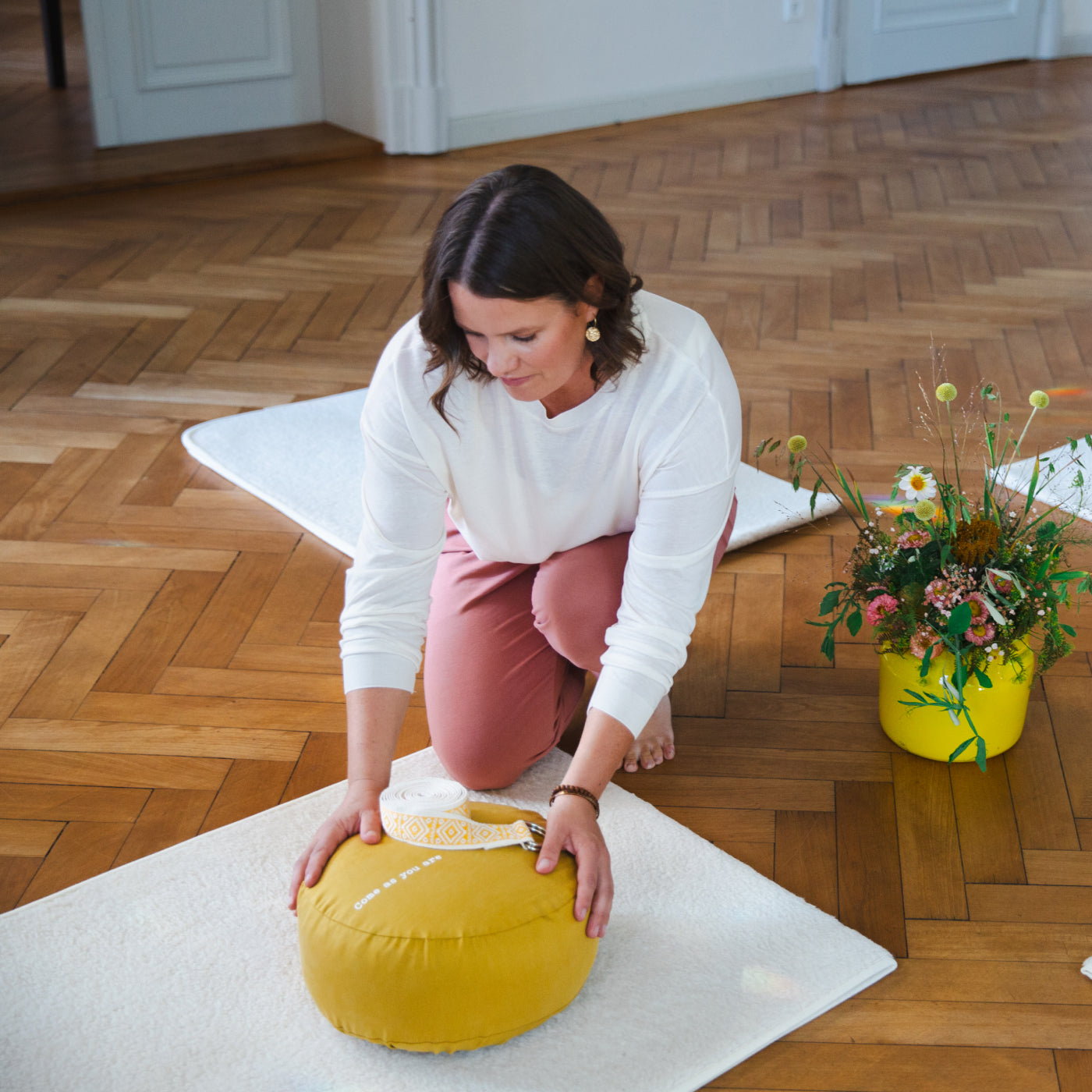 Eine Frau legt ein Meditationskissen und einen Yoga Gurt auf eine weiße Yogamatte.