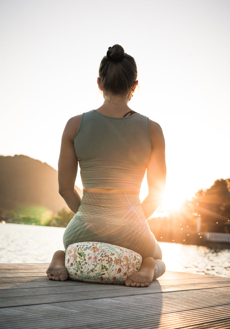 Eine Frau meditiert auf einem Goldwerk Schliersee Yogakissen im Sonnenuntergang vor einem Bergrücken.