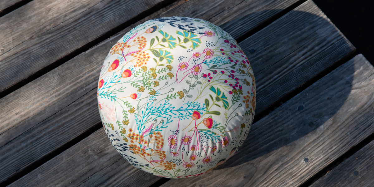 Ein weißes Meditationkissen mit bunten Blume bedruckt, liegt auf einem Holzsteg.