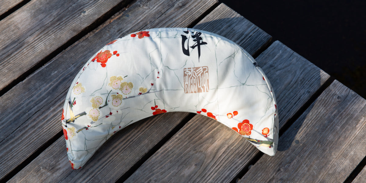 Ein weißes Yoga Halbmond Kissen mit roten Blume bedruckt, liegt auf einem Holzsteg.