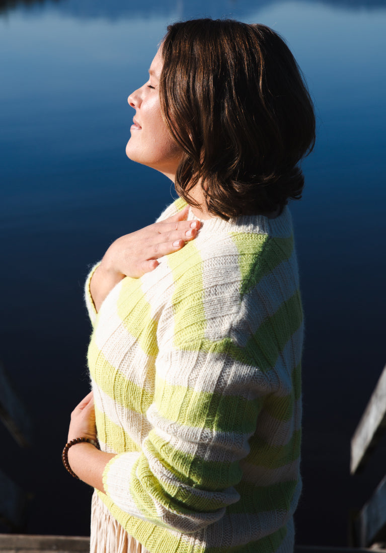 Mit geschlossenen Augen steht eine Frau am Wasser und hält ihr Gesicht der Sonne entgegen.