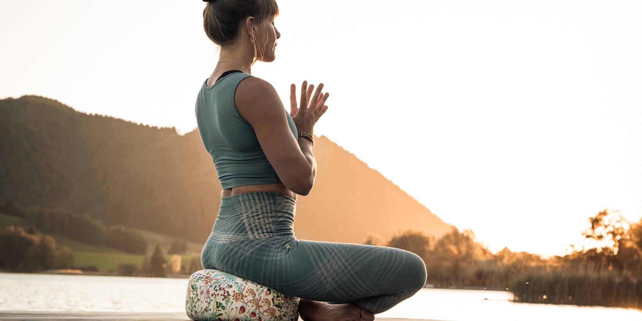Eine Frau meditiert auf einem Goldwerk Schliersee Meditationskissen im Sonnenuntergang vor einem Bergrücken.