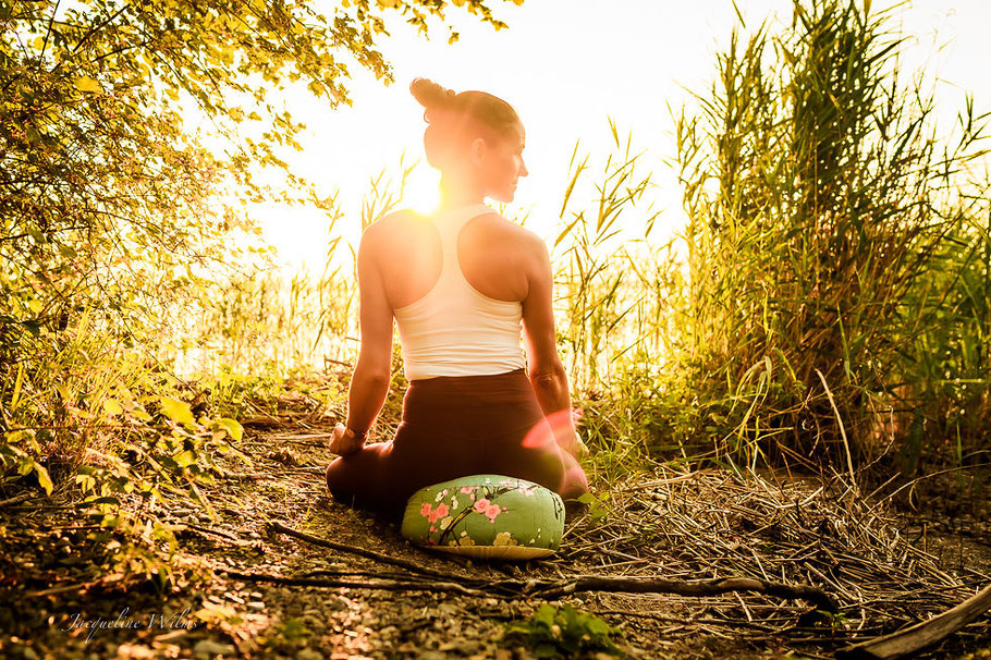 Im Gegenlicht zur Goldenen Stunde meditiert eine Frau auf einem bunten Meditationskissen am See.