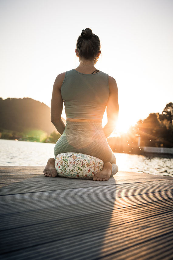Eine Frau von hinten auf einem Steg im Sonnenuntergang. Sie sitzt im Reitersitz auf einem Meditationskissen.