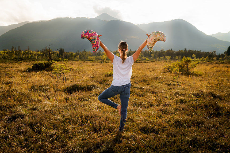 Eine Frau von hinten, die die Yogahaltung "Tadasana" übt. Sie hat die Arme nach oben gestreckt und hält jeweils ein Yoga Halbmondkissen in der Hand.