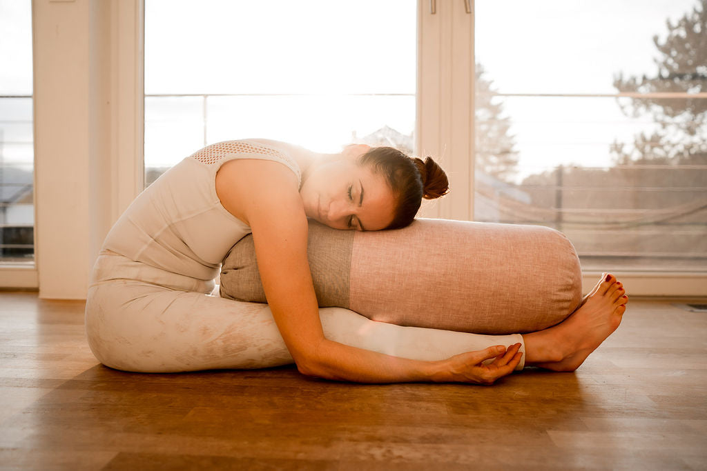 Frau übt eine Vorbeuge mit einem Yogabolster vor großen Fenstern im Gegenlicht.