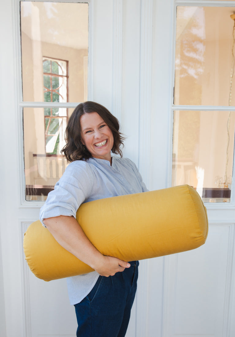 Eine Frau hält ein gelbes Yoga Bolster unter ihrem Arm und lächelt in die Kamera.