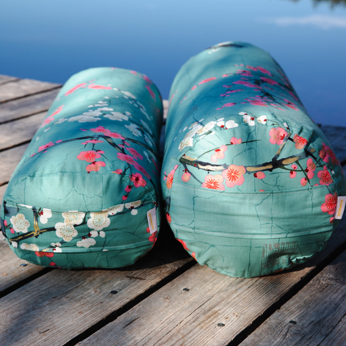 Zwei Türkise Bolster mit Kirschblüten Druck liegen auf einem Holzsteg an einem See.