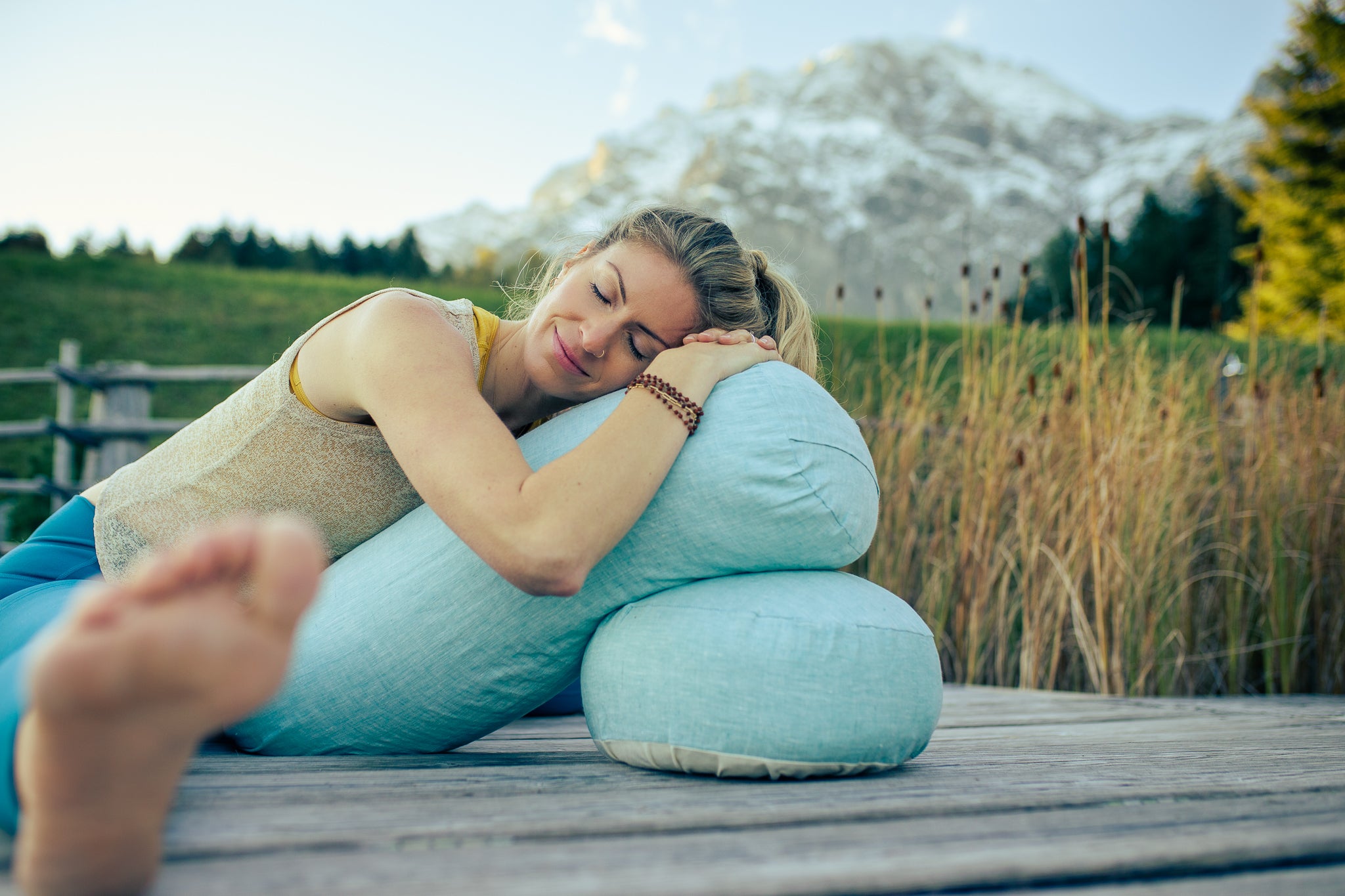 Frau übt Yin Yoga auf einem Yogabolster und einem Meditationskissen aus blauem Leinen vor einem Alpenpanorama