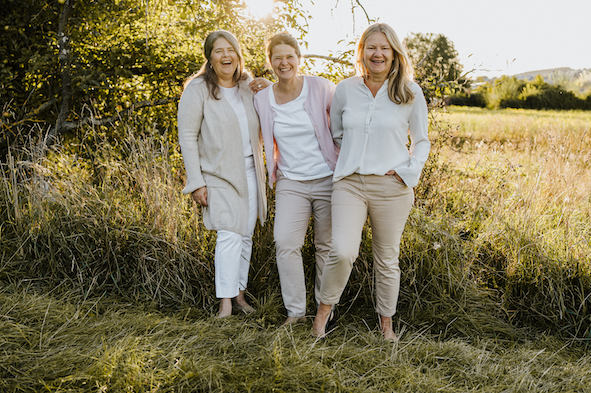 Drei Frauen halten sich im Arm und lachen in die Kamera vor Büschen und einer Wiese