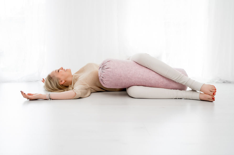 Eine blonde Frau übt die Yin Yoga Haltung "Gedrehte Wurzel" mit einem Yogabolster in einem weißen Raum.