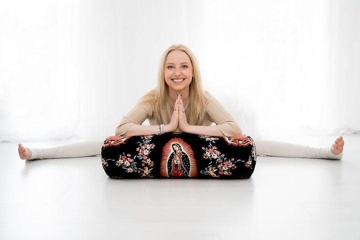 Eine lächelnde Frau sitzt in einer weiten Grätsche und stützt ihre Unterarme auf ein Yogabolster.