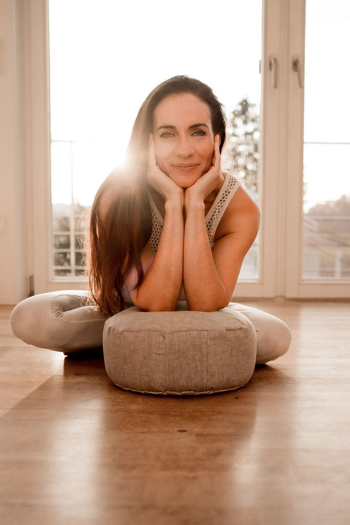 Frau mit langen Haaren sitzt mit überkreuzten Beinen hinter einem grauen Meditationskissen und stützt ihre Ellenbogen auf das Kissen. 