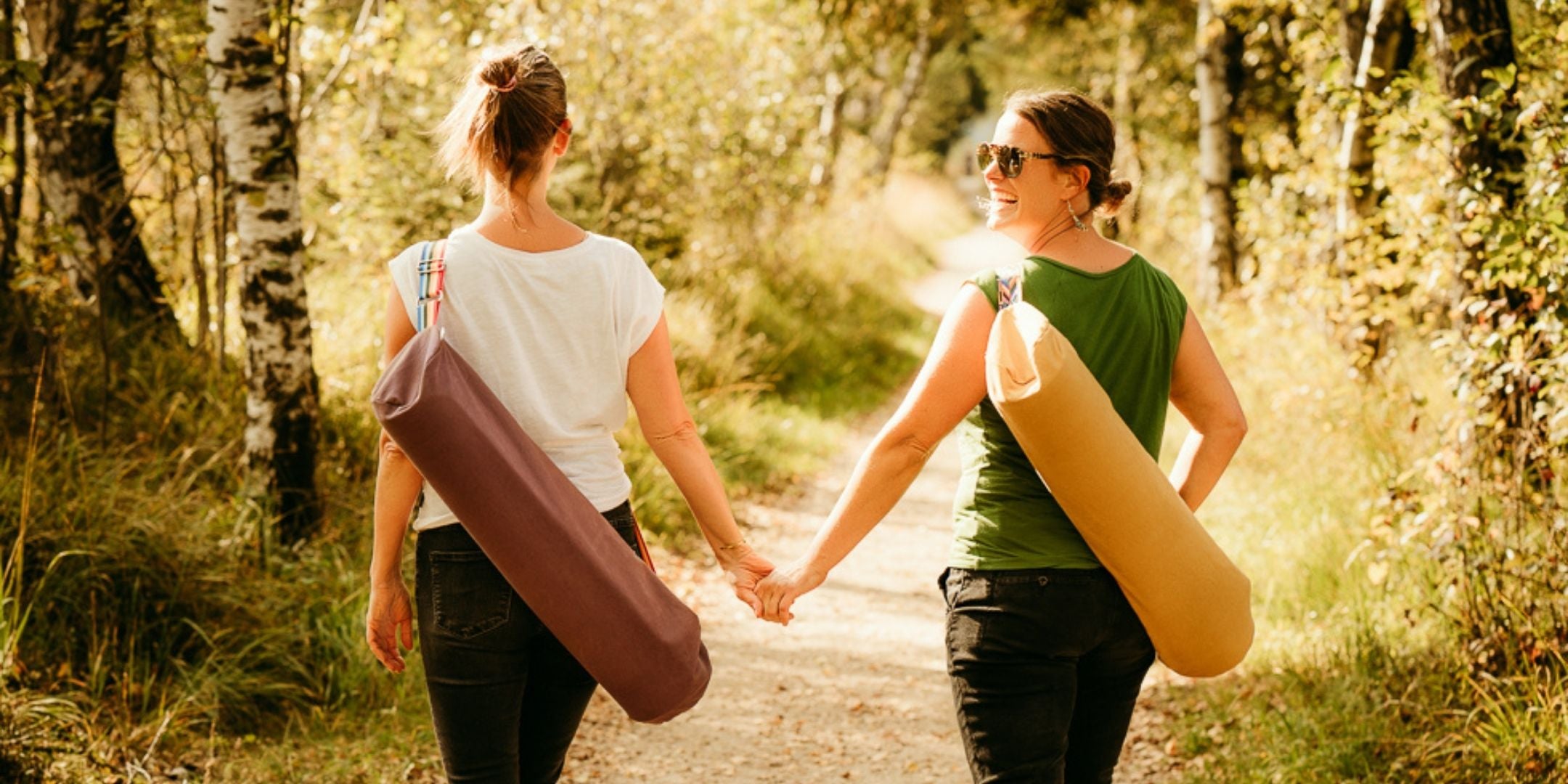 Zwei Freundinnen spazieren mit Yogataschen auf dem Rücken Hand in Hand auf einem Kiesweg in der Natur.